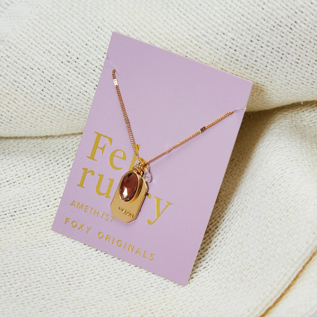A gold necklace with an amethyst stone pendant. The pendant also has the word "WISDOM" engraved on it. The necklace is displayed on a purple card with the month "February" and the brand name "Foxy Originals."