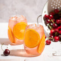 Two glasses of pink drinks with orange slices and a colander of cherries on a light surface.