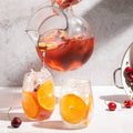 Person pouring iced tea into glasses with cherries and oranges on a table.
