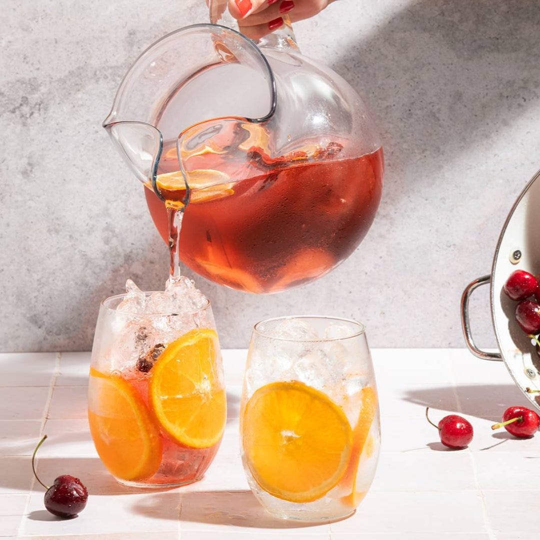 Person pouring iced tea into glasses with cherries and oranges on a table.