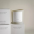 A beige glass jar candle with a white label. The label reads "NORTH ROSE" and "FIRESIDE."
