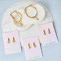 A flat lay of various gold jewelry pieces displayed on a white marble surface. The items include necklaces with star and sun pendants, earrings with star and heart charms, and a layered bracelet set. The brand name "Foxy Originals" is visible on the packaging cards.