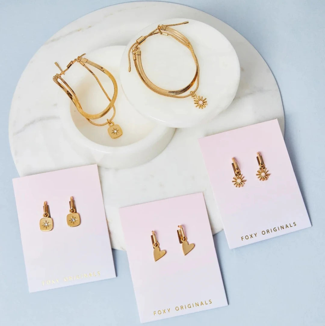 A flat lay of various gold jewelry pieces displayed on a white marble surface. The items include necklaces with star and sun pendants, earrings with star and heart charms, and a layered bracelet set. The brand name "Foxy Originals" is visible on the packaging cards.