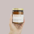 Hand holding a jar of 'Frasier Fir' scented candle against a plain background
