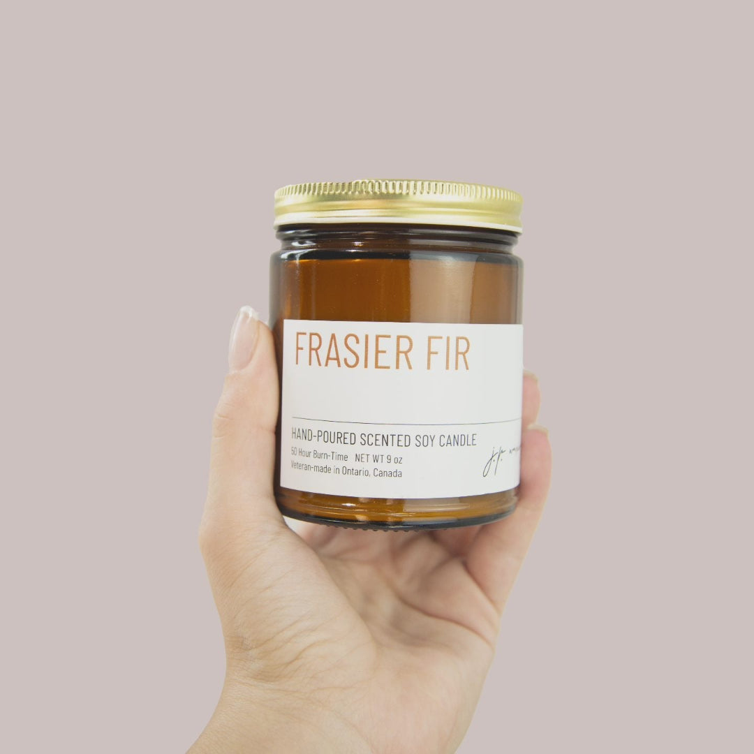 Hand holding a jar of 'Frasier Fir' scented candle against a plain background