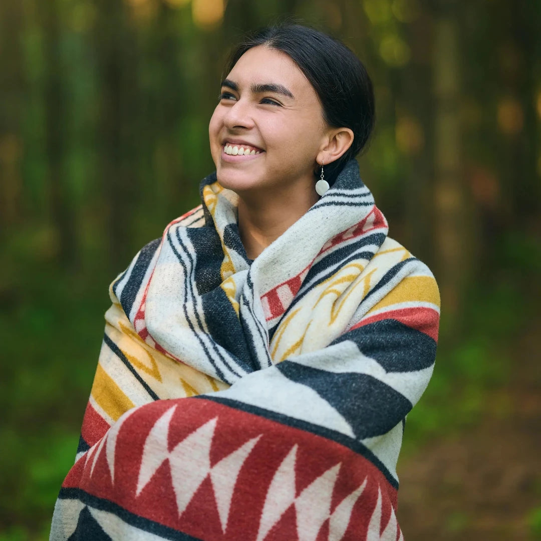 The Generations Reversible Throw Blanket. The blanket features a bold, geometric pattern with stripes, triangles, and diamond shapes in shades of red, black, white, and yellow. It is draped over a branch and has a fringed edge.