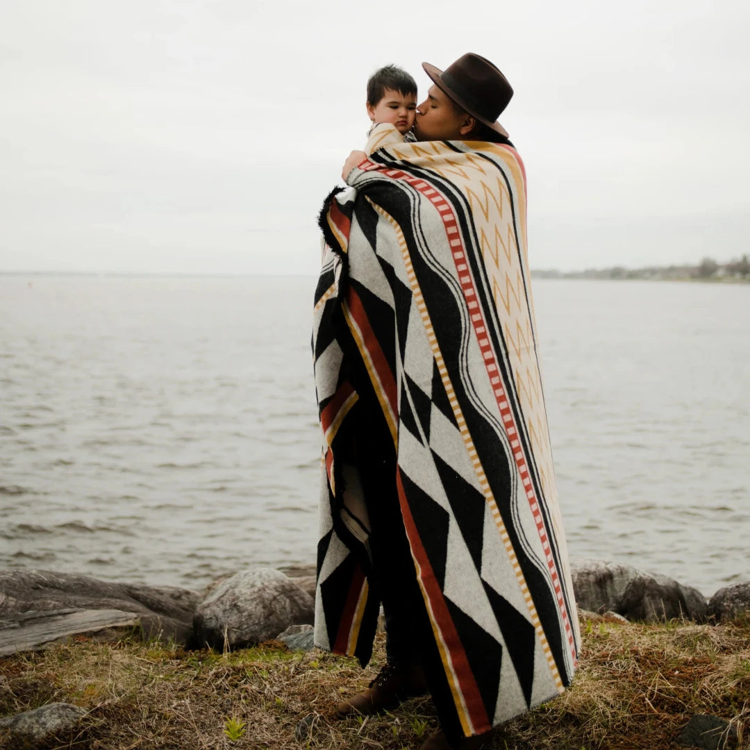 The Generations Reversible Throw Blanket. The blanket features a bold, geometric pattern with stripes, triangles, and diamond shapes in shades of red, black, white, and yellow. It is draped over a branch and has a fringed edge.