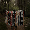 The Generations Reversible Throw Blanket. The blanket features a bold, geometric pattern with stripes, triangles, and diamond shapes in shades of red, black, white, and yellow. It is draped over a branch and has a fringed edge.