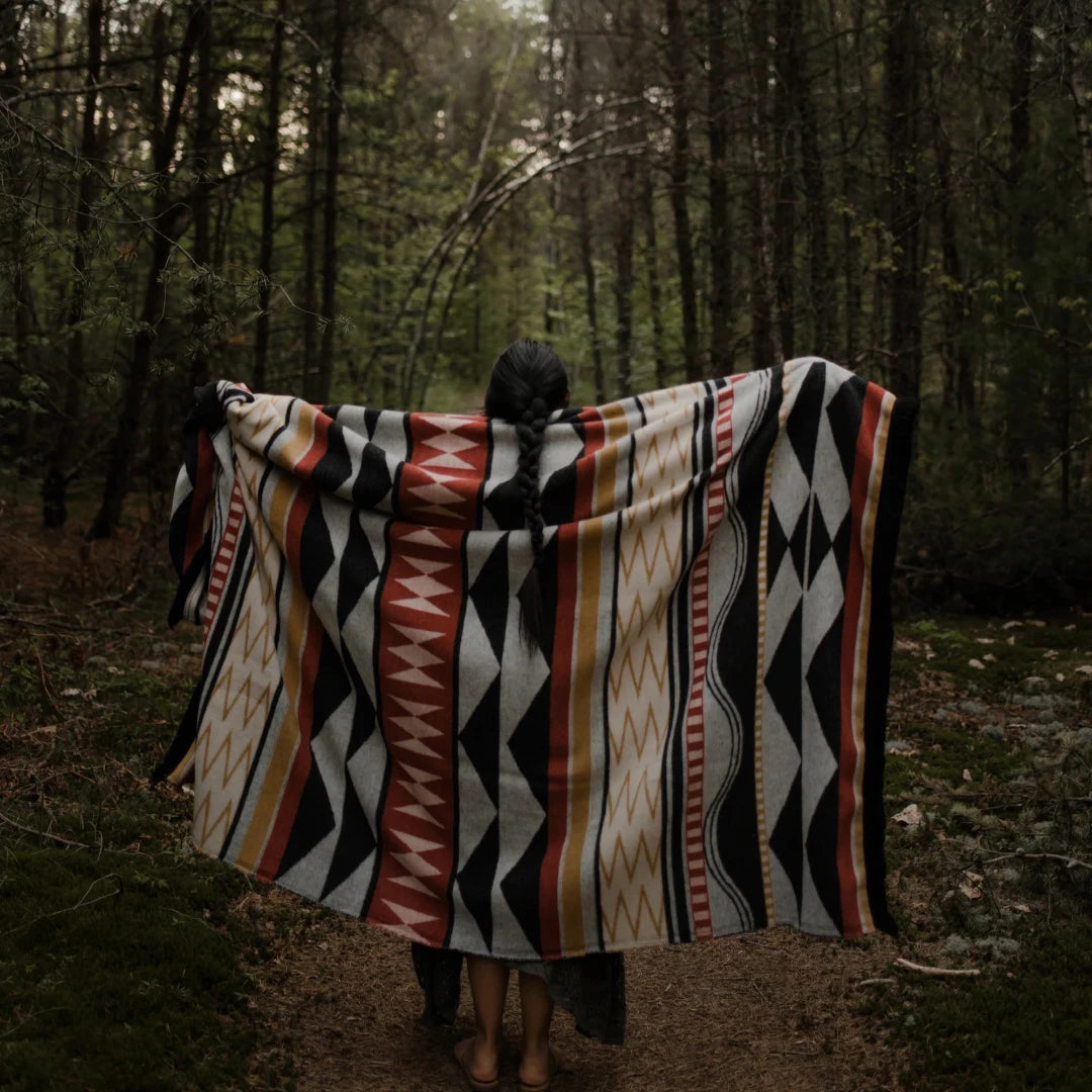 The Generations Reversible Throw Blanket. The blanket features a bold, geometric pattern with stripes, triangles, and diamond shapes in shades of red, black, white, and yellow. It is draped over a branch and has a fringed edge.