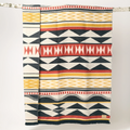 The Generations Reversible Throw Blanket. The blanket features a bold, geometric pattern with stripes, triangles, and diamond shapes in shades of red, black, white, and yellow. It is draped over a branch and has a fringed edge.