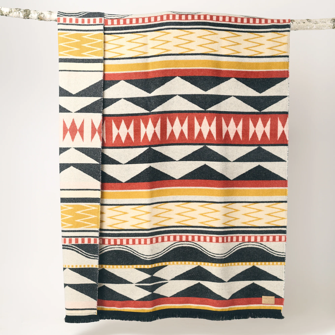 The Generations Reversible Throw Blanket. The blanket features a bold, geometric pattern with stripes, triangles, and diamond shapes in shades of red, black, white, and yellow. It is draped over a branch and has a fringed edge.