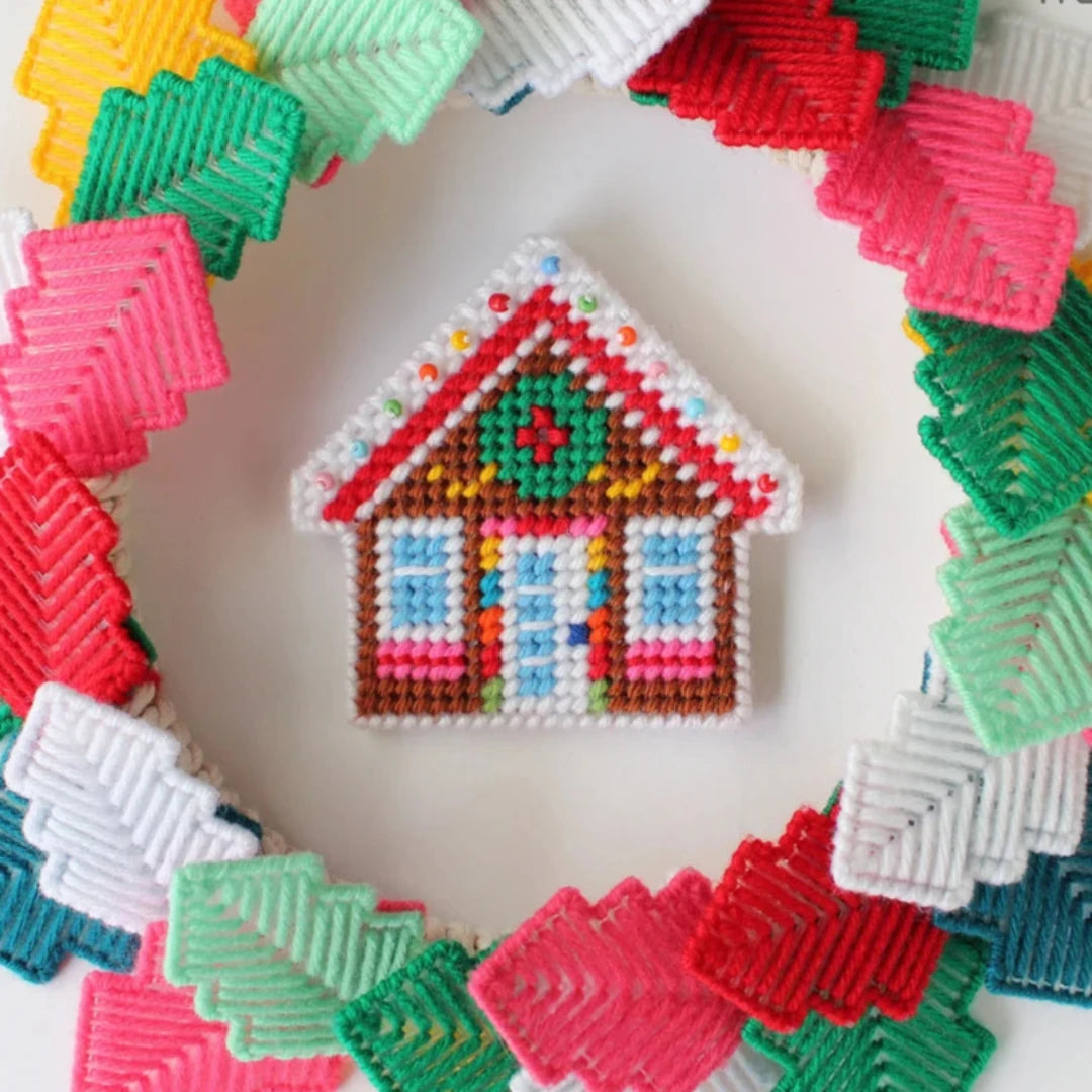 a colorful plastic canvas gingerbread house ornament. The house is decorated with red and white candy cane trim, a green wreath, and blue and white windows.