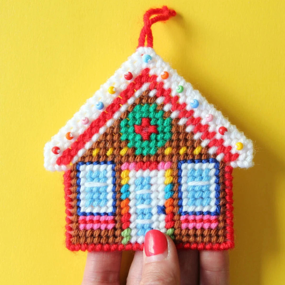 a colorful plastic canvas gingerbread house ornament. The house is decorated with red and white candy cane trim, a green wreath, and blue and white windows.