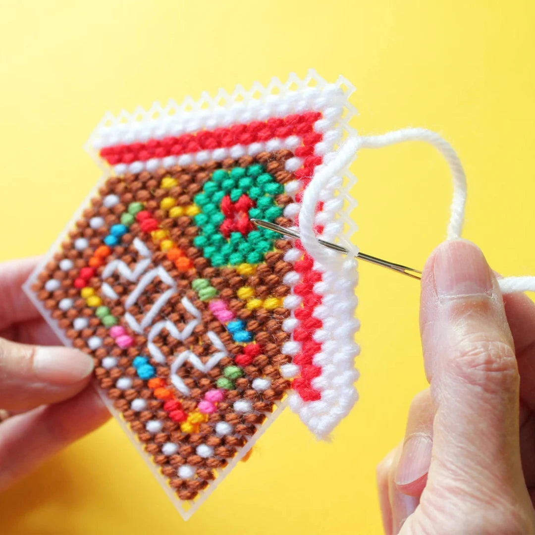 a colorful plastic canvas gingerbread house ornament. The house is decorated with red and white candy cane trim, a green wreath, and blue and white windows.