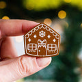 A brown gingerbread house pin on a blue background. The gingerbread house has white icing details, including a bow, windows, and a door. There are also snowflakes on the roof.