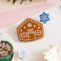 A brown gingerbread house pin on a blue background. The gingerbread house has white icing details, including a bow, windows, and a door. There are also snowflakes on the roof.