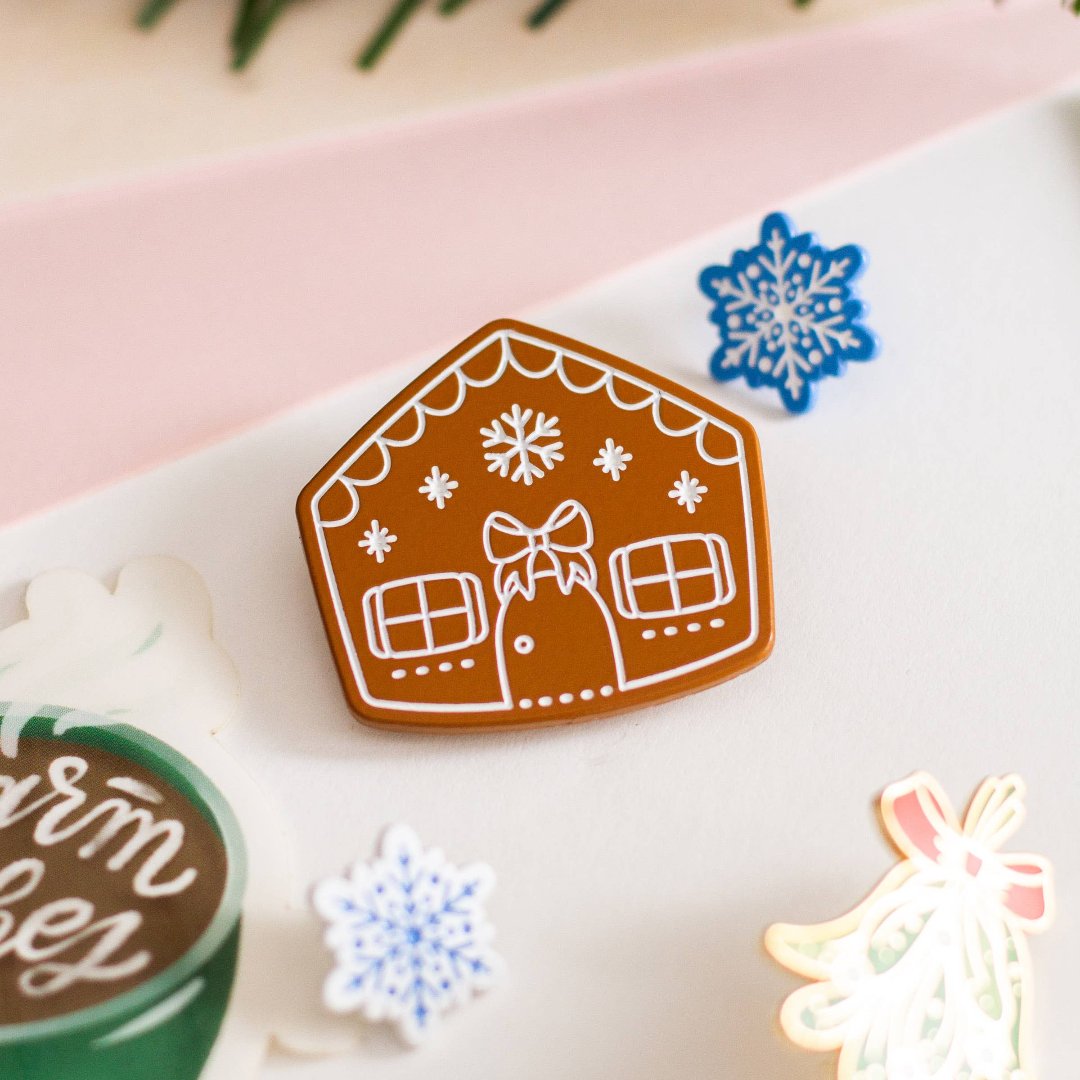 A brown gingerbread house pin on a blue background. The gingerbread house has white icing details, including a bow, windows, and a door. There are also snowflakes on the roof.