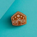 A brown gingerbread house pin on a blue background. The gingerbread house has white icing details, including a bow, windows, and a door. There are also snowflakes on the roof.