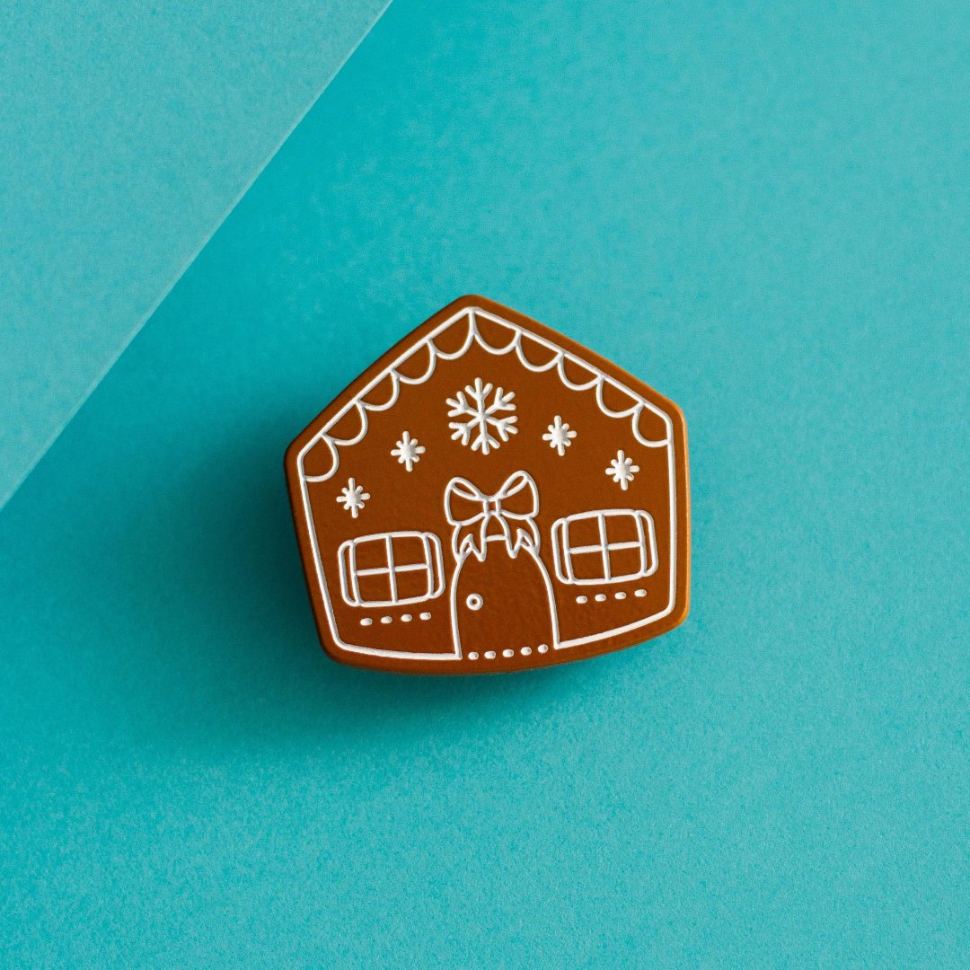 A brown gingerbread house pin on a blue background. The gingerbread house has white icing details, including a bow, windows, and a door. There are also snowflakes on the roof.