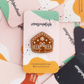 A brown gingerbread house pin on a blue background. The gingerbread house has white icing details, including a bow, windows, and a door. There are also snowflakes on the roof.