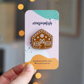 A brown gingerbread house pin on a blue background. The gingerbread house has white icing details, including a bow, windows, and a door. There are also snowflakes on the roof.