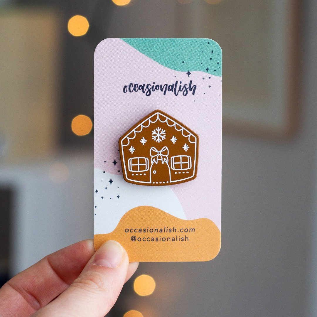 A brown gingerbread house pin on a blue background. The gingerbread house has white icing details, including a bow, windows, and a door. There are also snowflakes on the roof.