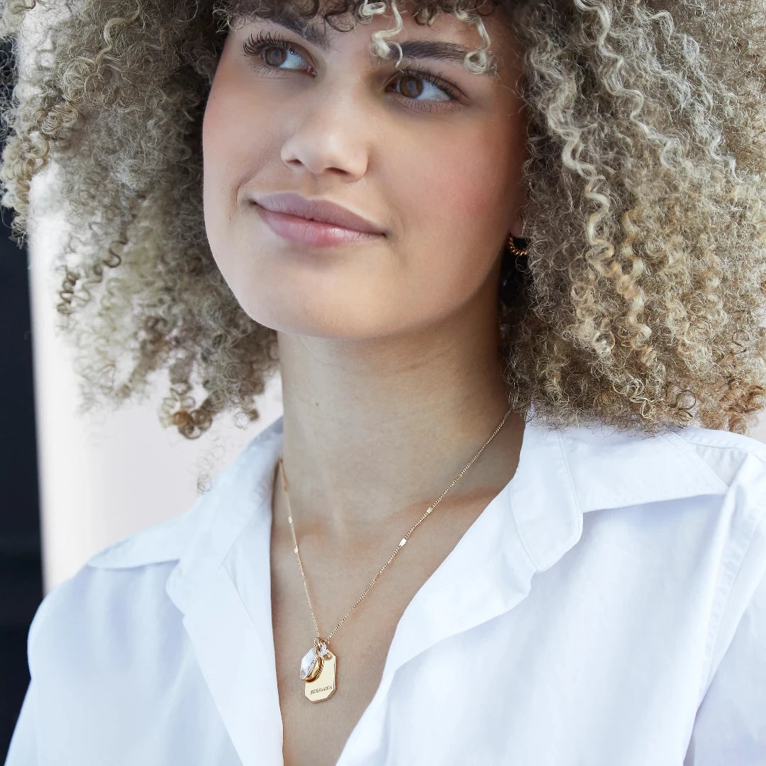 A gold necklace with a quartz stone pendant. The pendant also has the word "STRENGTH" engraved on it. The necklace is displayed on a pink card with the month "April" and the brand name "Foxy Originals."
