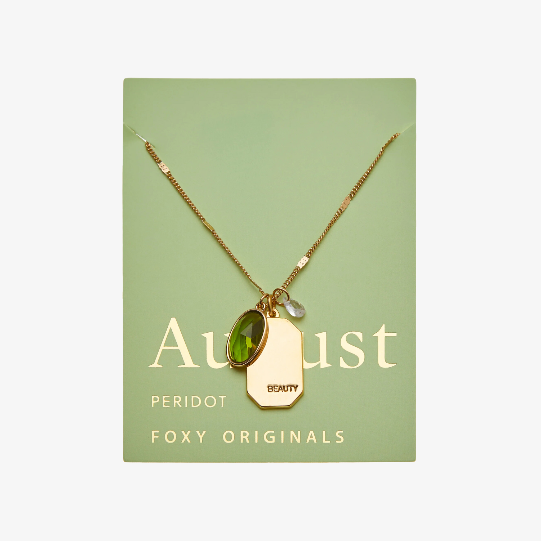 A gold necklace with a peridot stone pendant. The pendant also has the word "BEAUTY" engraved on it. The necklace is displayed on a green card with the month "August" and the brand name "Foxy Originals."