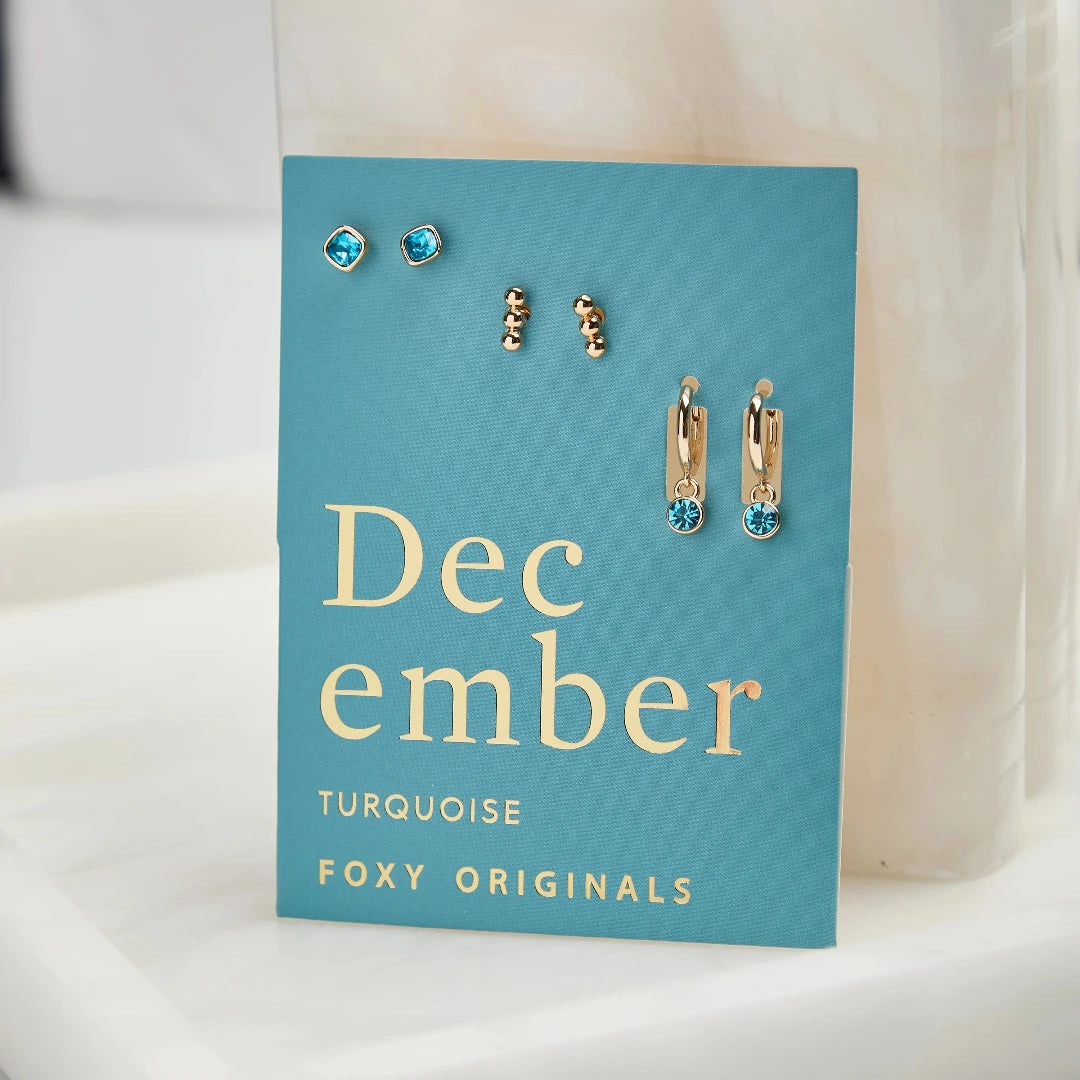 A set of three pairs of turquoise earrings displayed on a blue card. The set includes a pair of stud earrings, a pair of bar earrings with small turquoise beads, and a pair of hoop earrings with dangling turquoise stones. The card has the month "December" and the brand name "Foxy Originals."