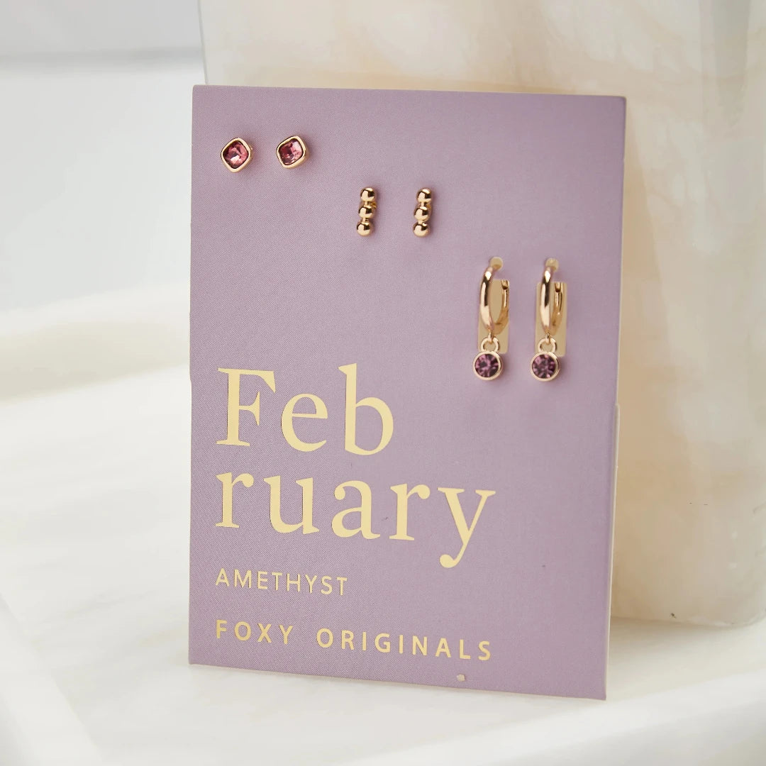 A set of three pairs of amethyst earrings displayed on a purple card. The set includes a pair of stud earrings, a pair of bar earrings with small amethyst beads, and a pair of hoop earrings with dangling amethyst stones. The card has the month "February" and the brand name "Foxy Originals."