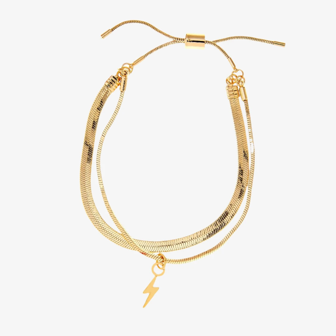 A set of two gold bracelets, one layered over the other, with a lightning bolt charm hanging from the bottom. The bracelets are displayed on a pink card with the brand name "Foxy Originals."