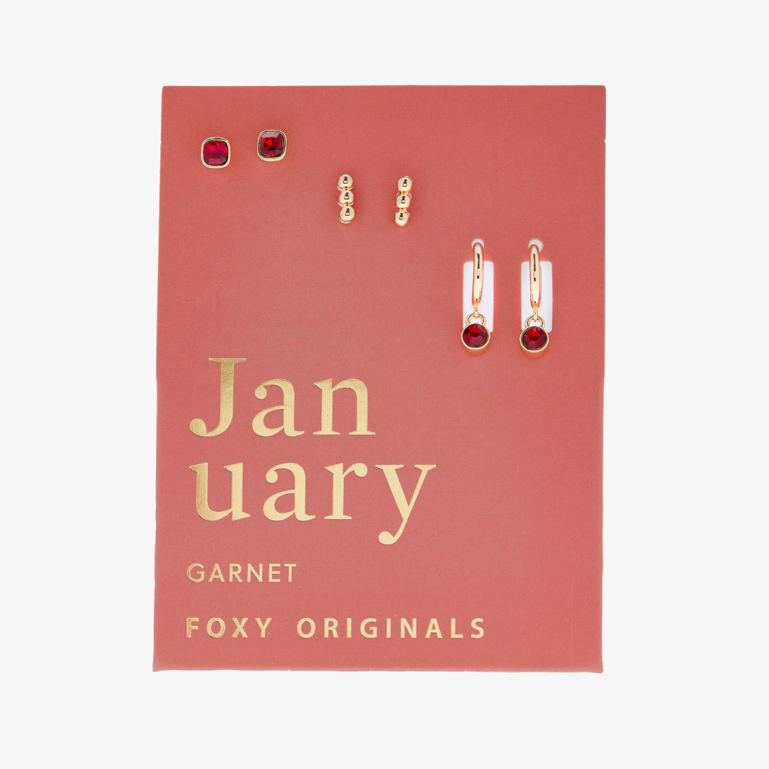 A set of three pairs of garnet earrings displayed on a red card. The set includes a pair of stud earrings, a pair of bar earrings with small garnet beads, and a pair of hoop earrings with dangling garnet stones. The card has the month "January" and the brand name "Foxy Originals."