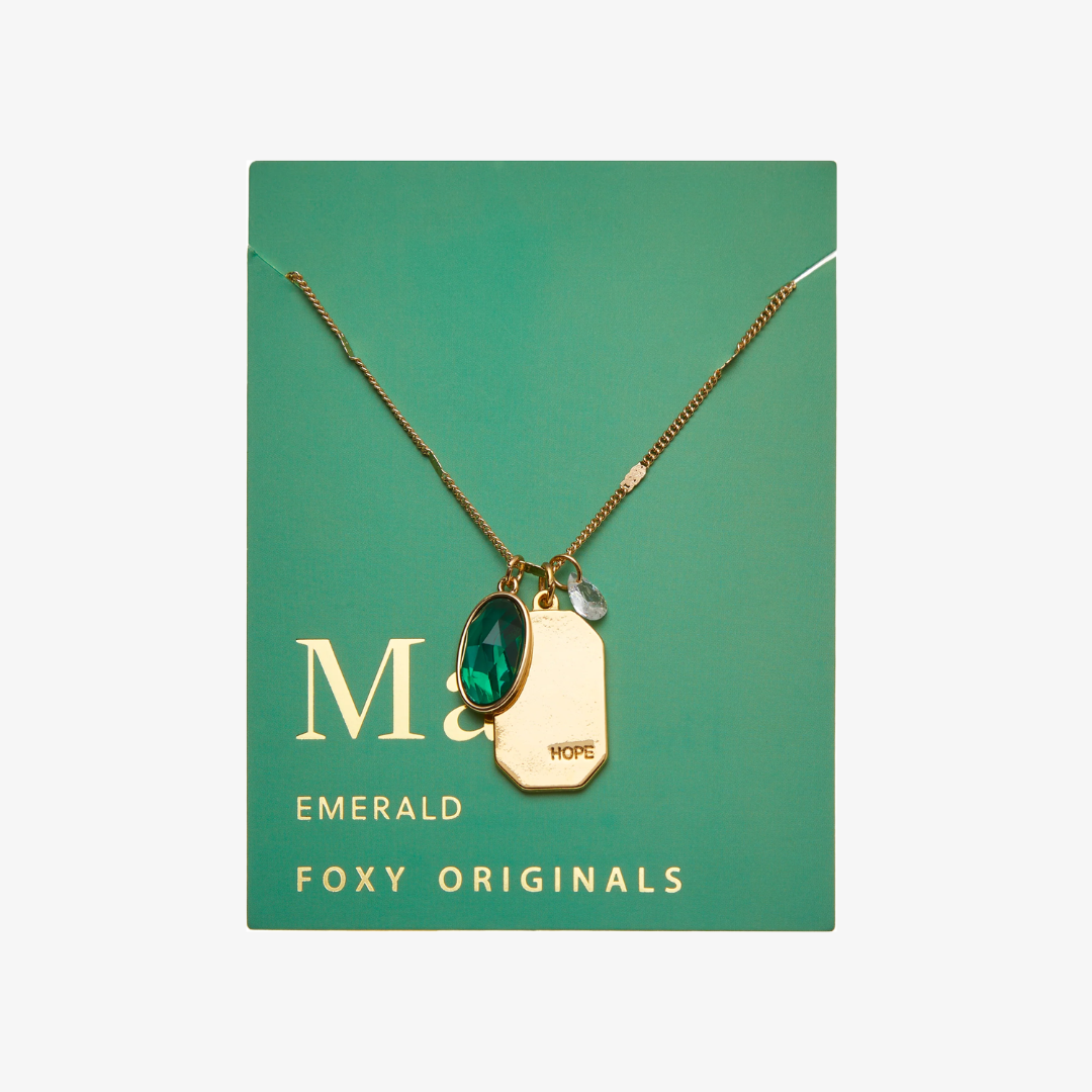 A gold necklace with an emerald stone pendant. The pendant also has the word "HOPE" engraved on it. The necklace is displayed on a green card with the month "May" and the brand name "Foxy Originals."
