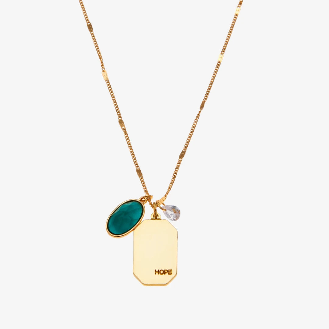 A gold necklace with an emerald stone pendant. The pendant also has the word "HOPE" engraved on it. The necklace is displayed on a green card with the month "May" and the brand name "Foxy Originals."
