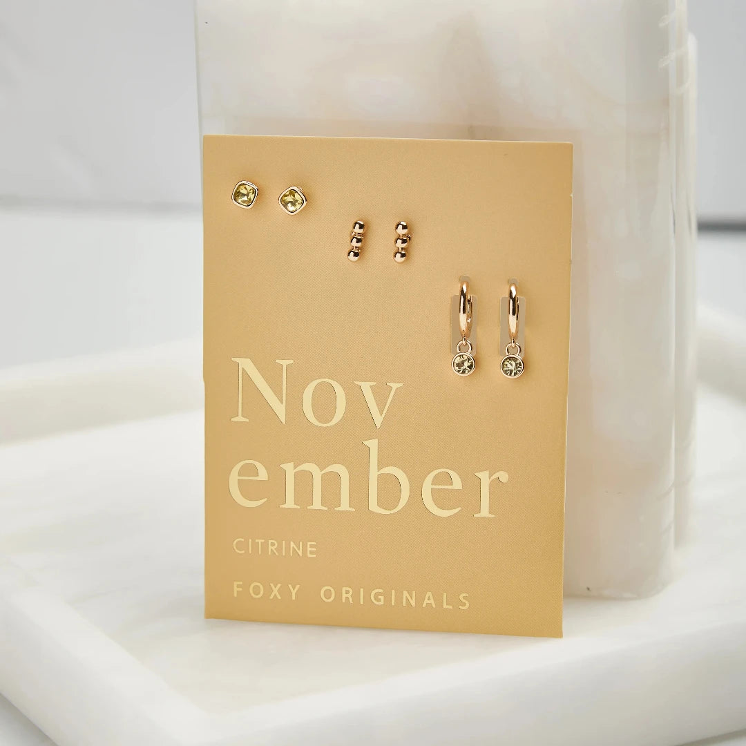 A set of three pairs of citrine earrings displayed on a yellow card. The set includes a pair of stud earrings, a pair of bar earrings with small citrine beads, and a pair of hoop earrings with dangling citrine stones. The card has the month "November" and the brand name "Foxy Originals."