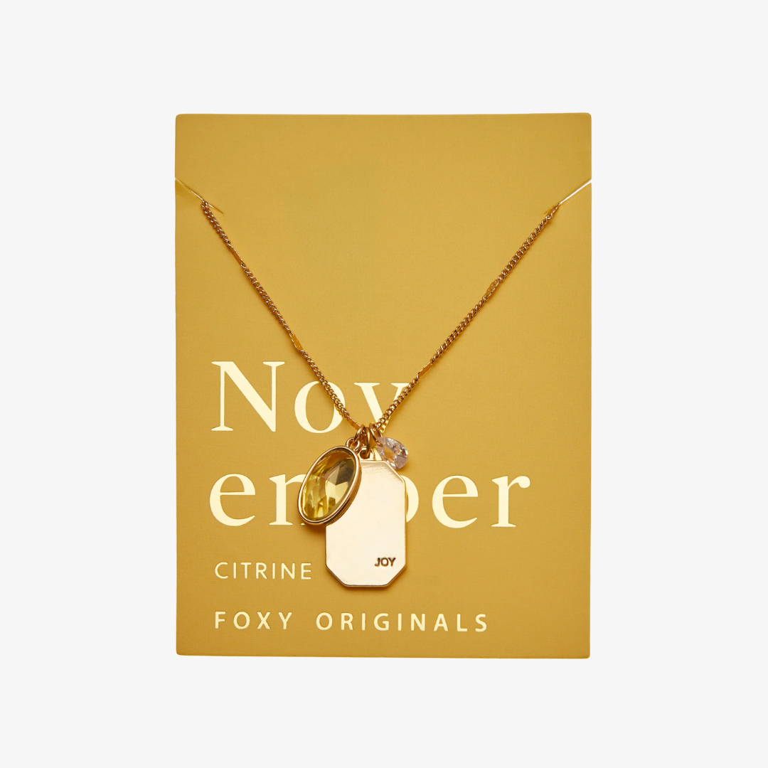 A gold necklace with a citrine stone pendant. The pendant also has the word "JOY" engraved on it. The necklace is displayed on a yellow card with the month "November" and the brand name "Foxy Originals."