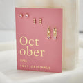 A set of three pairs of opal earrings displayed on a pink card. The set includes a pair of stud earrings, a pair of bar earrings with small opal beads, and a pair of hoop earrings with dangling opal stones. The card has the month "October" and the brand name "Foxy Originals."