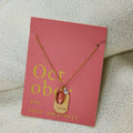 A gold necklace with an opal stone pendant. The pendant also has the word "HEALING" engraved on it. The necklace is displayed on a pink card with the month "October" and the brand name "Foxy Originals."