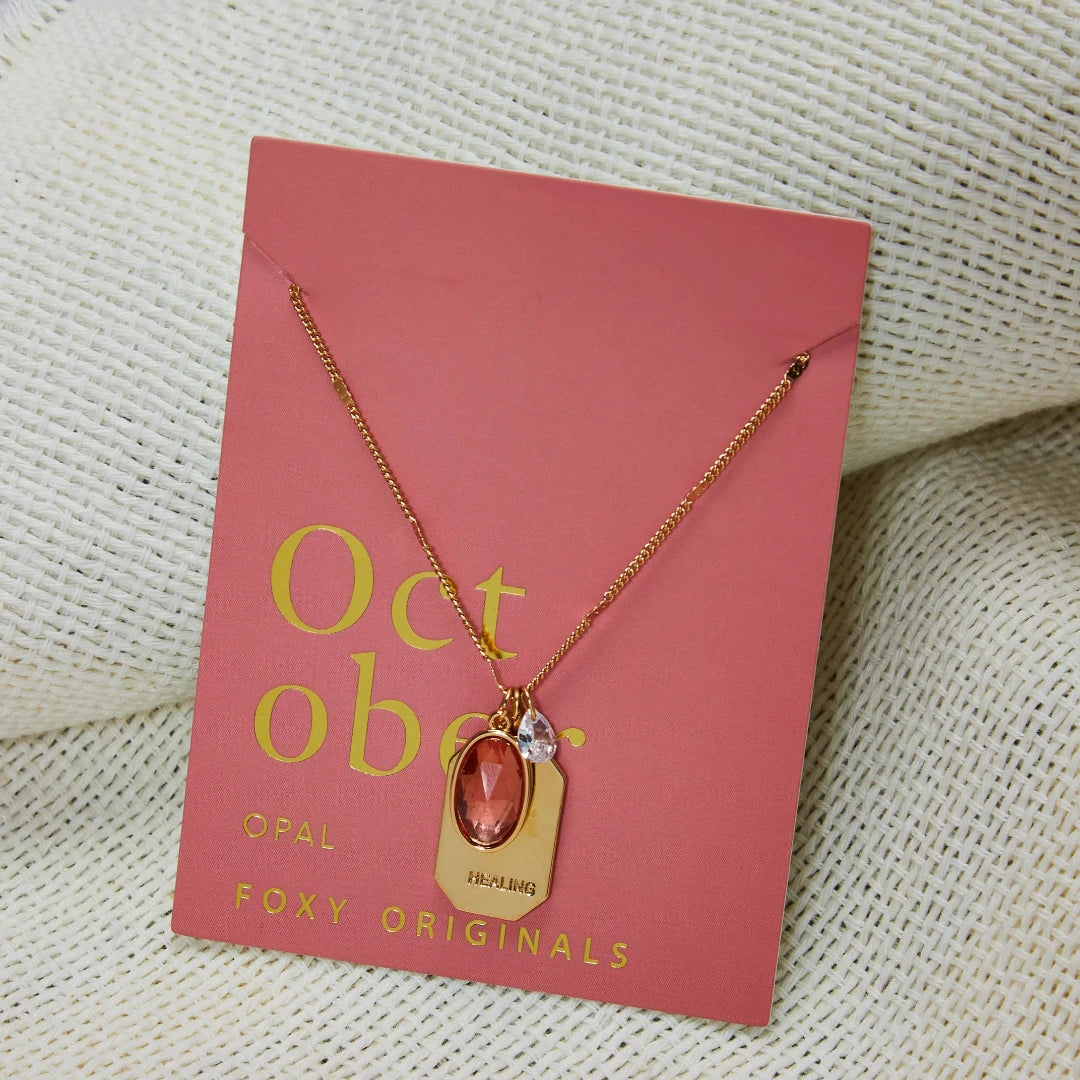 A gold necklace with an opal stone pendant. The pendant also has the word "HEALING" engraved on it. The necklace is displayed on a pink card with the month "October" and the brand name "Foxy Originals."