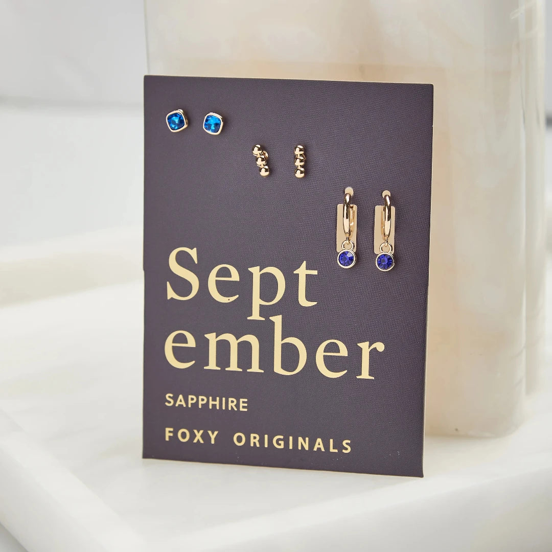 A set of three pairs of sapphire earrings displayed on a blue card. The set includes a pair of stud earrings, a pair of bar earrings with small sapphire beads, and a pair of hoop earrings with dangling sapphire stones. The card has the month "September" and the brand name "Foxy Originals."