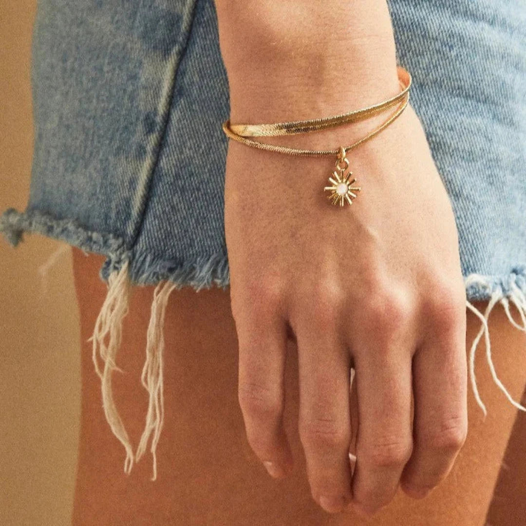 A set of two gold bracelets, one layered over the other, with a sun charm hanging from the bottom. The bracelets are displayed on a pink card with the brand name "Foxy Originals."