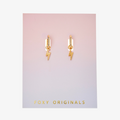 A pair of gold earrings with lightning bolt charms on a pink background. The earrings are displayed on a card with the brand name "Foxy Originals."