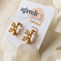 A pair of Agaveh Girl gold Forget Me Not Earrings, featuring a small, gold flower design with a beige center, displayed on a branded card with the text 'agaveh girl,' against a textured background with white floral accents. The earrings are dainty and floral, available in Ottawa, Ontario, Canada.