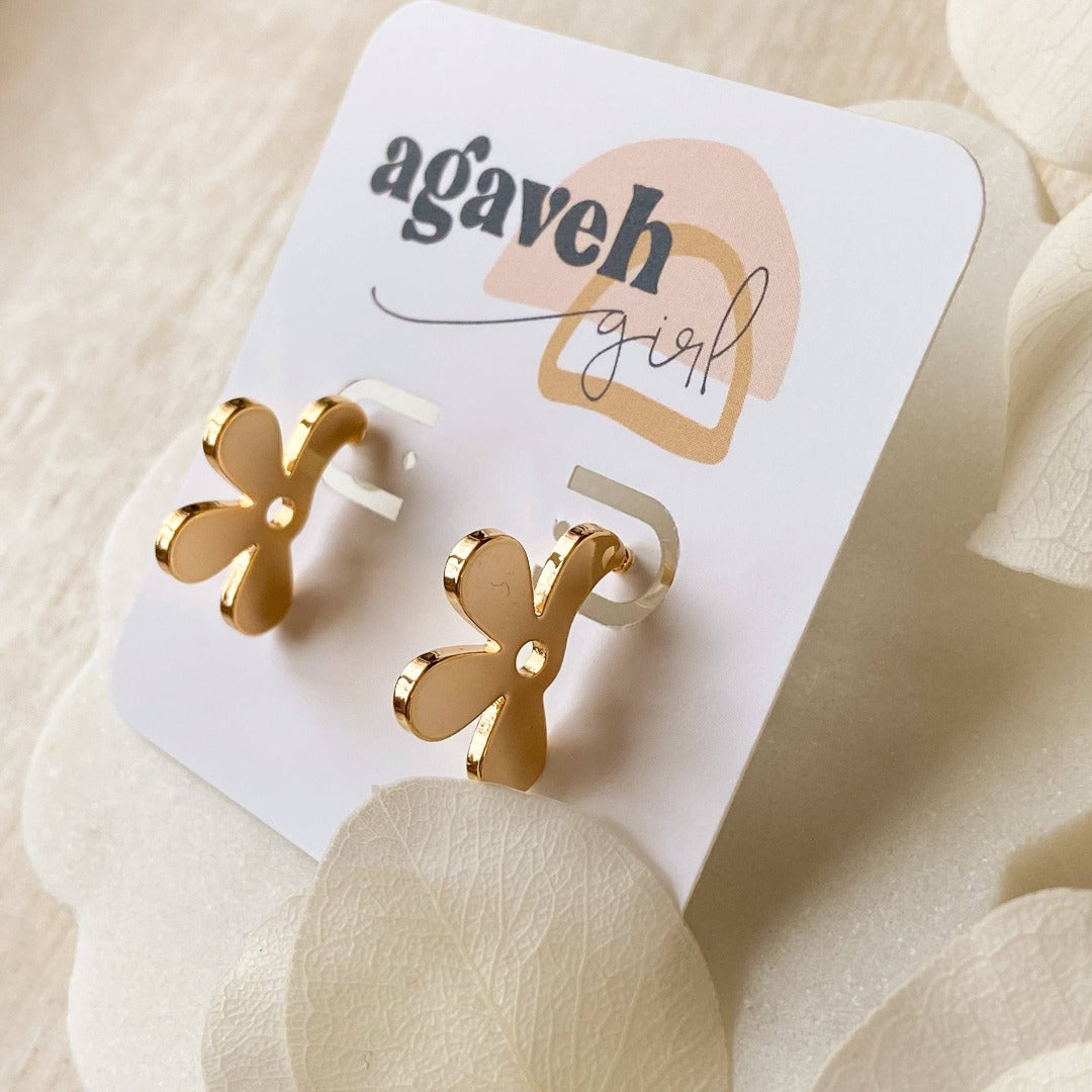 A pair of Agaveh Girl gold Forget Me Not Earrings, featuring a small, gold flower design with a beige center, displayed on a branded card with the text 'agaveh girl,' against a textured background with white floral accents. The earrings are dainty and floral, available in Ottawa, Ontario, Canada.