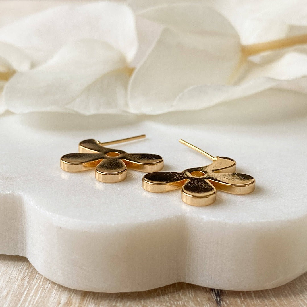 A pair of Agaveh Girl gold Forget Me Not Earrings, featuring a small, gold flower design with a beige center, displayed on a branded card with the text 'agaveh girl,' against a textured background with white floral accents. The earrings are dainty and floral, available in Ottawa, Ontario, Canada.