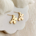 A pair of Agaveh Girl gold Forget Me Not Earrings, featuring a small, gold flower design with a beige center, displayed on a branded card with the text 'agaveh girl,' against a textured background with white floral accents. The earrings are dainty and floral, available in Ottawa, Ontario, Canada.