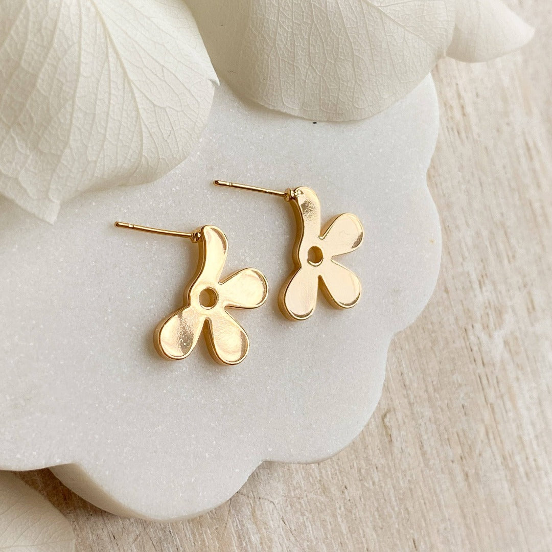A pair of Agaveh Girl gold Forget Me Not Earrings, featuring a small, gold flower design with a beige center, displayed on a branded card with the text 'agaveh girl,' against a textured background with white floral accents. The earrings are dainty and floral, available in Ottawa, Ontario, Canada.