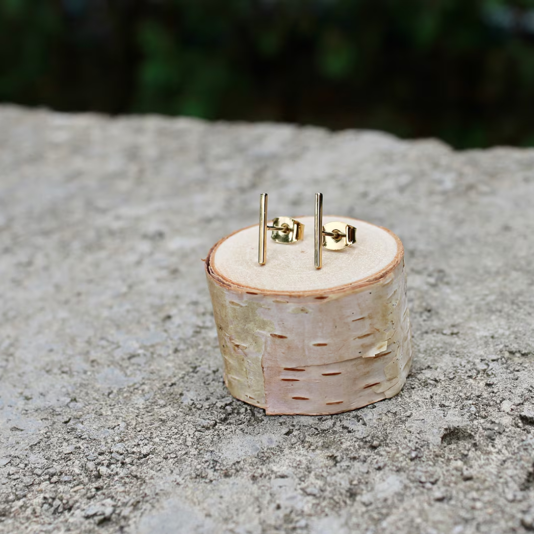 A pair of gold bar stud earrings resting on a small wooden disc. The earrings are simple and minimalist, with a single straight bar for each earring.