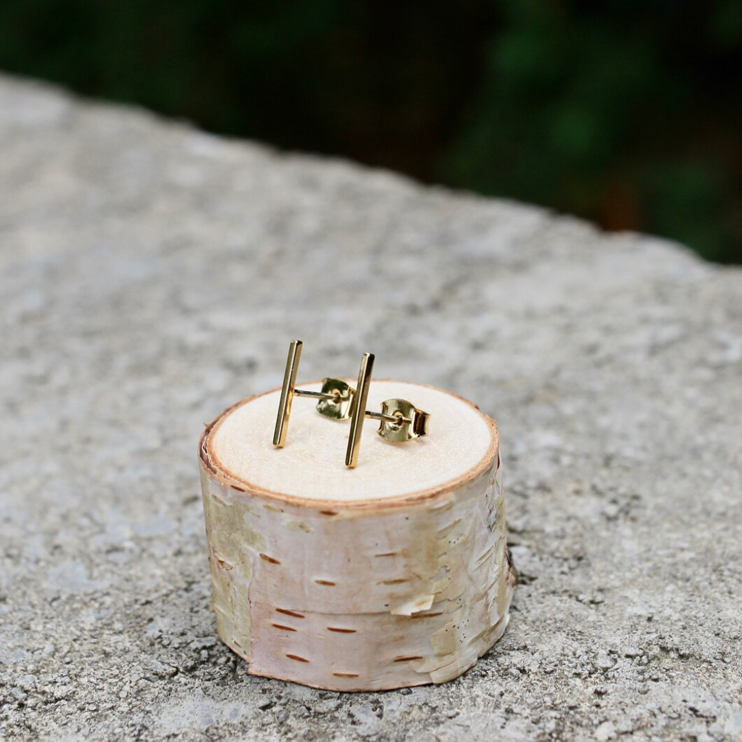 A pair of gold bar stud earrings resting on a small wooden disc. The earrings are simple and minimalist, with a single straight bar for each earring.