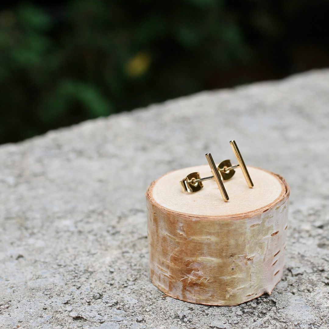A pair of gold bar stud earrings resting on a small wooden disc. The earrings are simple and minimalist, with a single straight bar for each earring.